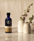 On a marble table, next to two plants, is a blue 100ml Neal's Yard Remedies Aromatic Massage Oil bottle.
