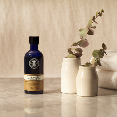 On a marble table, next to two plants, is a blue 100ml Neal's Yard Remedies Aromatic Massage Oil bottle.
