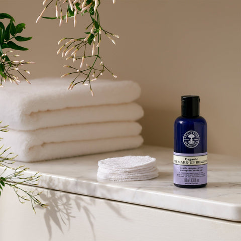 Neal's Yard Remedies' Organic Eye Make Up Remover blue bottom on a granite table besides 2 stacked bath towels.